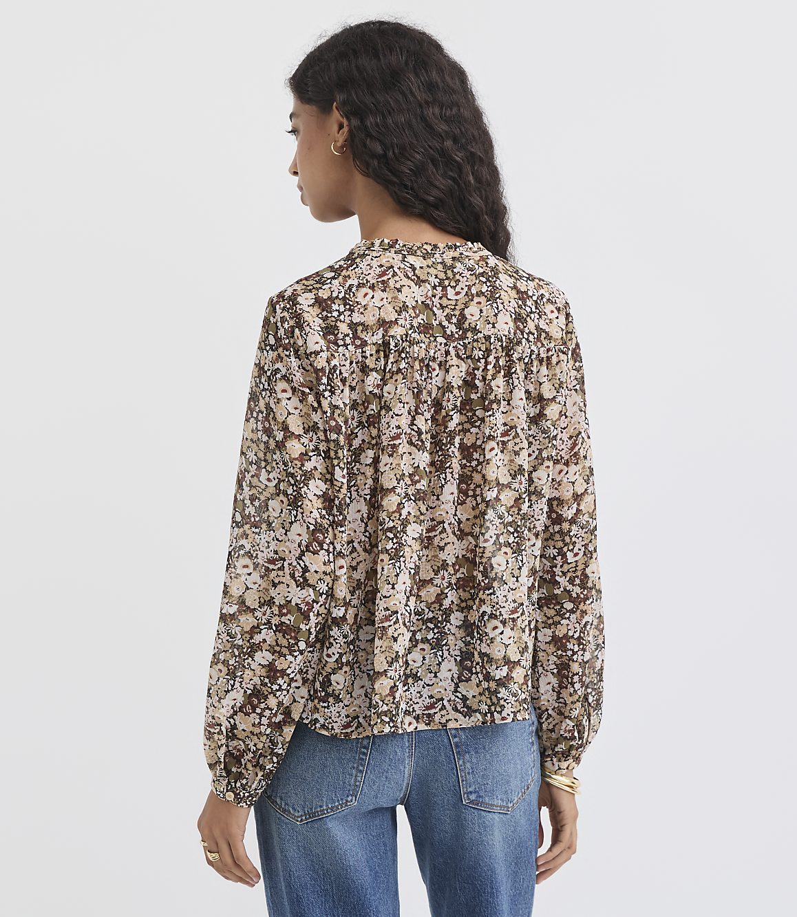 A woman in a floral blouse and jeans stands against a plain, light gray background.