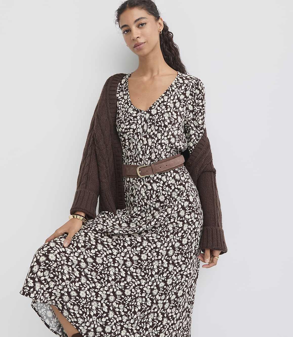 A woman in a floral dress and brown cardigan stands against a plain white background.