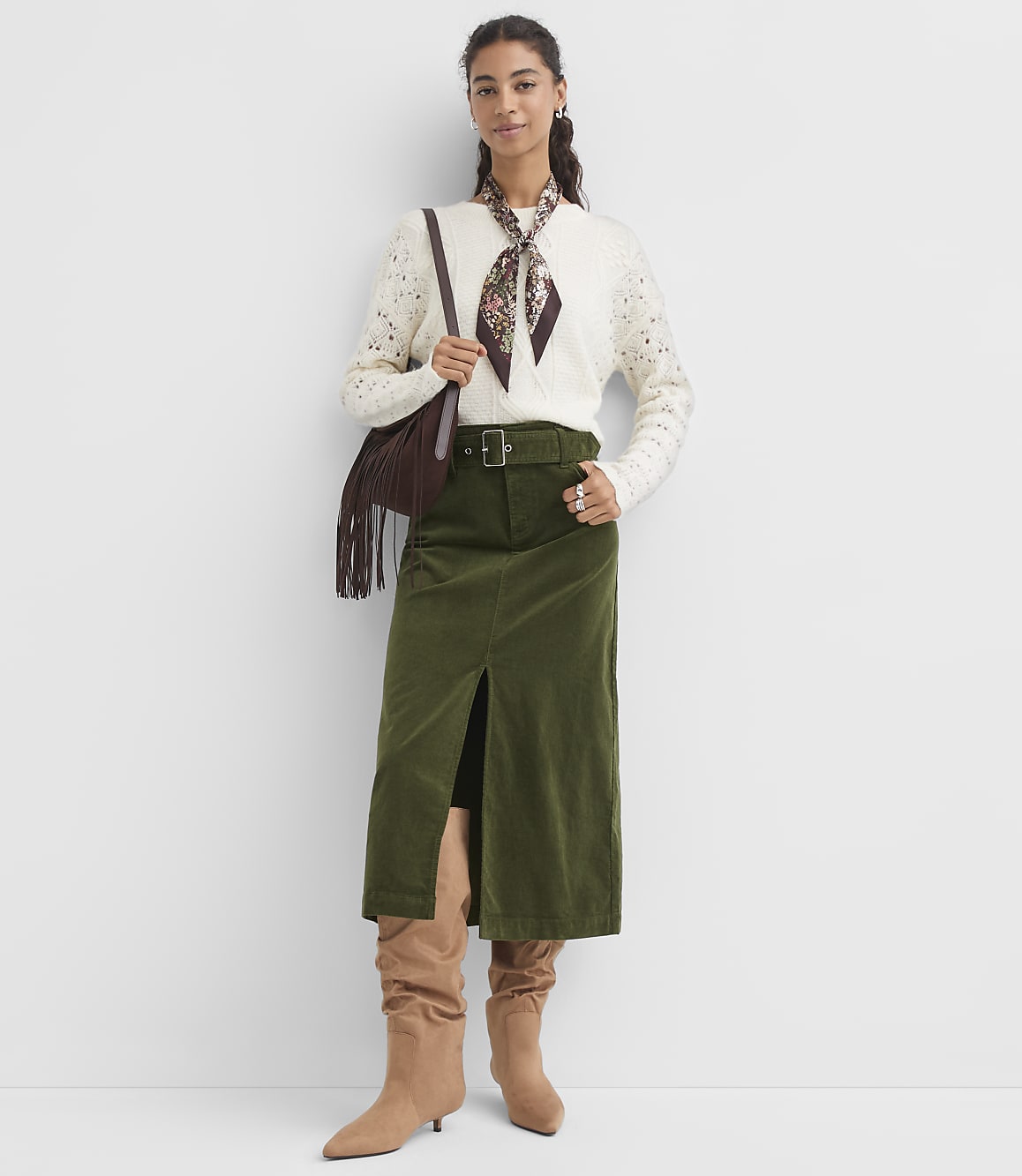 A woman in a cream sweater and olive green skirt stands against a plain white background, accessorized with a patterned scarf and a fringed bag.