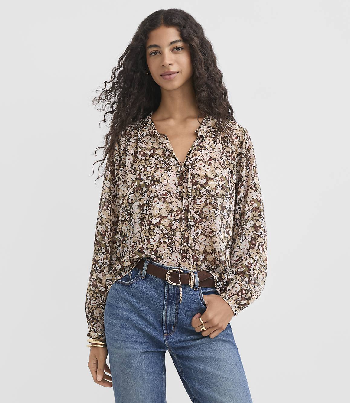 A woman in a floral blouse and jeans stands against a plain background.