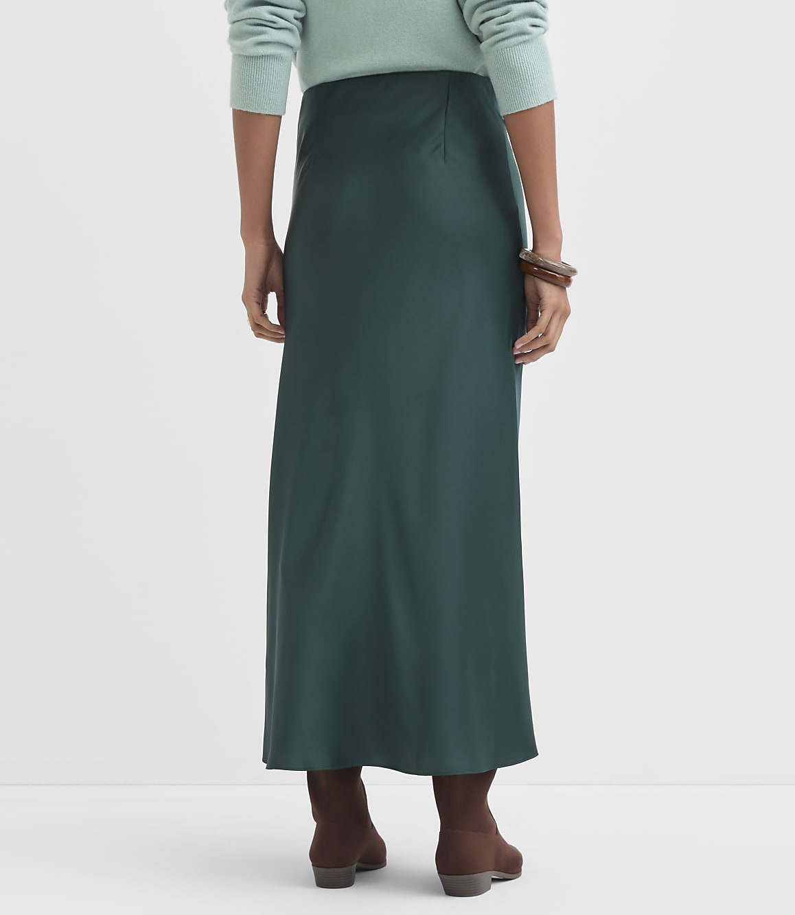 A person wearing a teal sweater and a dark green satin skirt stands against a plain white background, with brown boots visible at their feet.