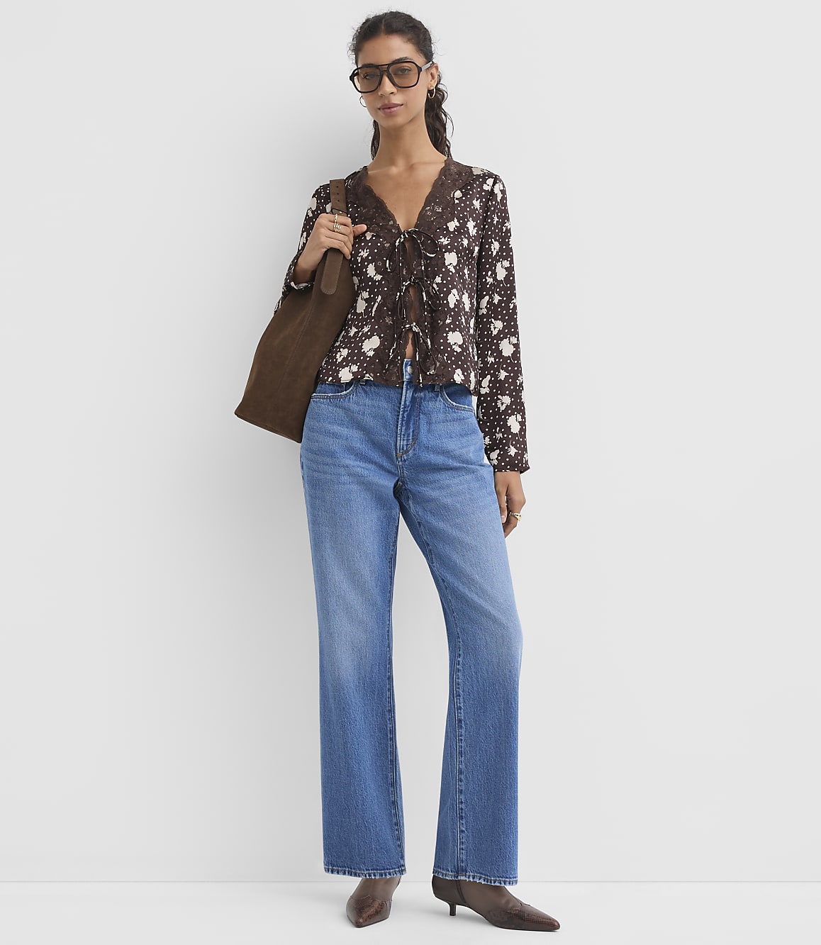 A woman in a floral blouse and jeans stands against a plain white background, carrying a brown tote bag.