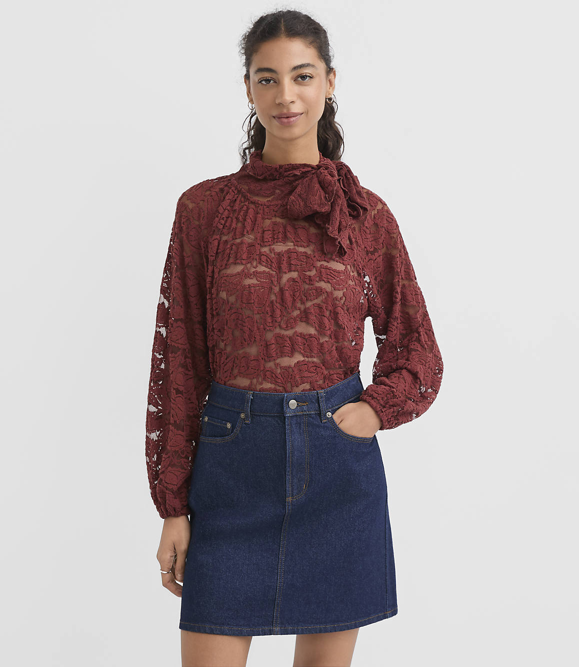 A woman in a maroon lace blouse and denim skirt stands against a plain gray background.