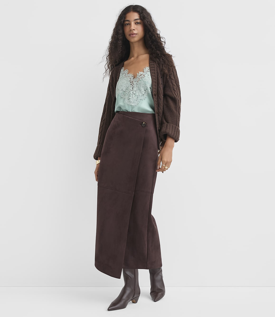 A woman in a brown cardigan, mint camisole, and brown suede skirt stands against a plain white background.