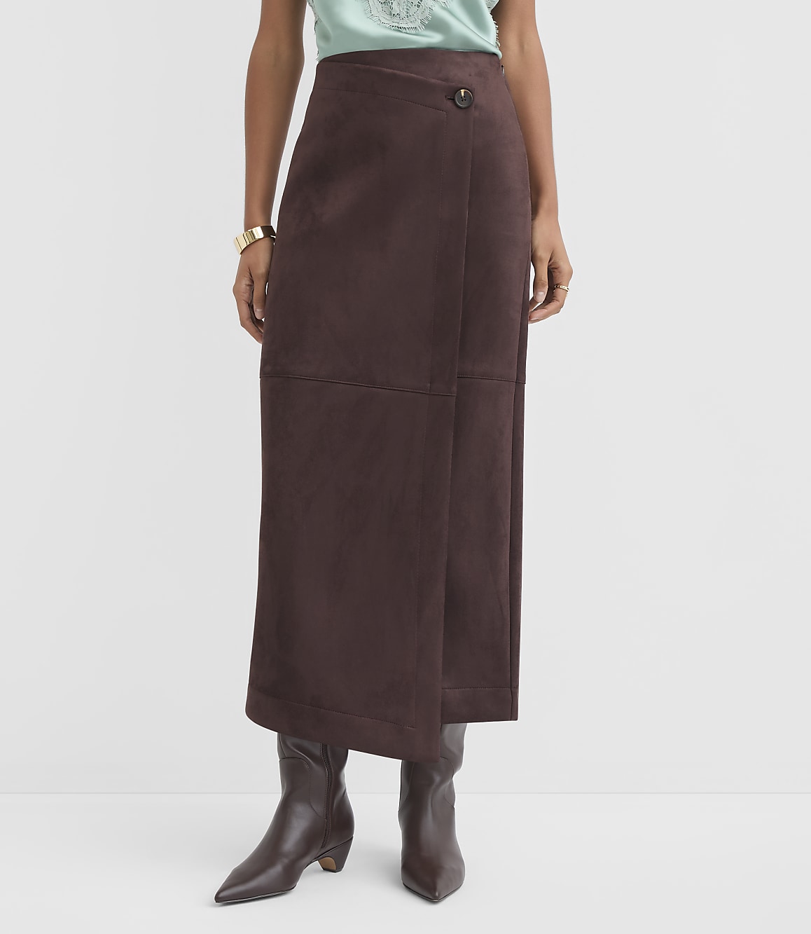 A person stands in the foreground wearing a brown wrap skirt and dark boots against a plain gray background.