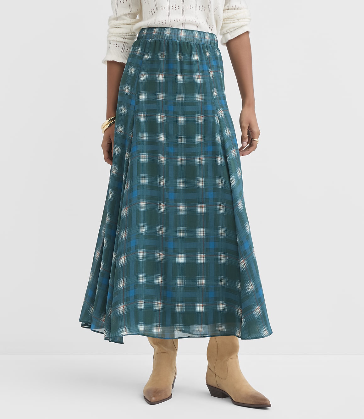 A woman in a teal plaid maxi skirt and tan boots stands against a plain white background.