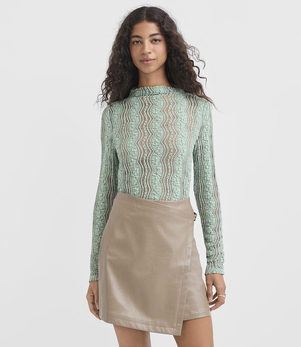 A woman in a green lace top and a tan faux leather skirt stands against a plain, light gray background.