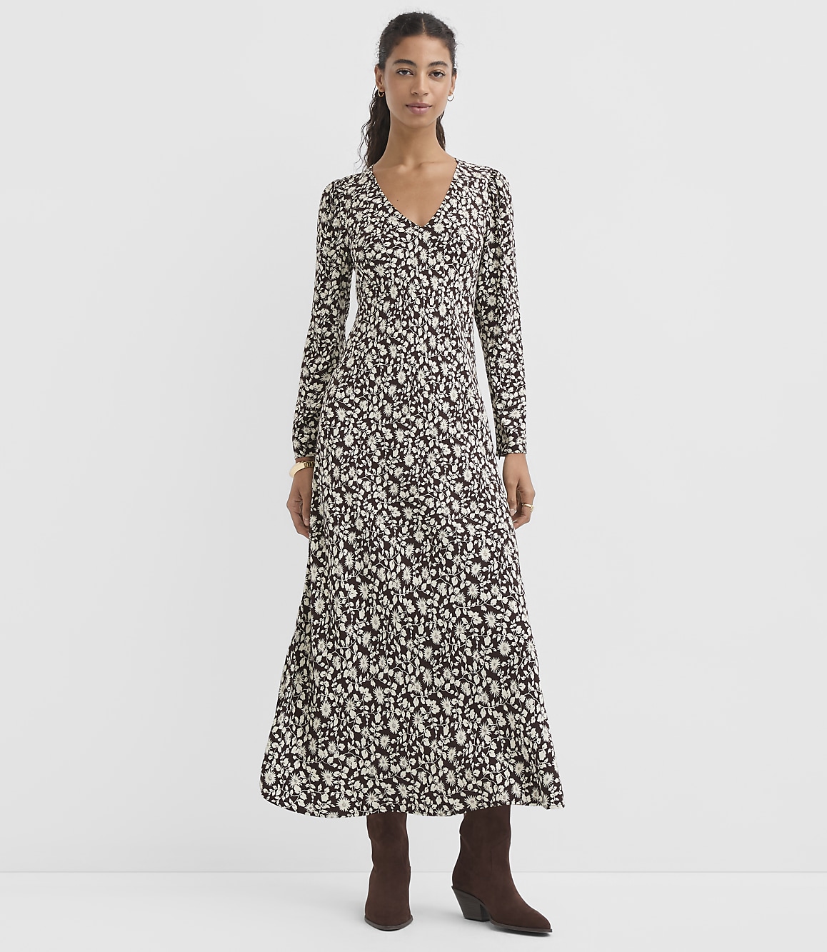 A woman in a long, patterned dress and brown boots stands against a plain, light gray background.