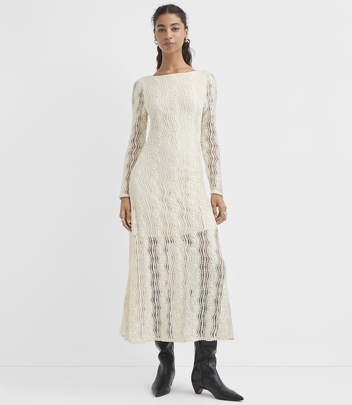 A woman in a cream-colored, textured, long-sleeved dress and black boots stands against a plain white background.