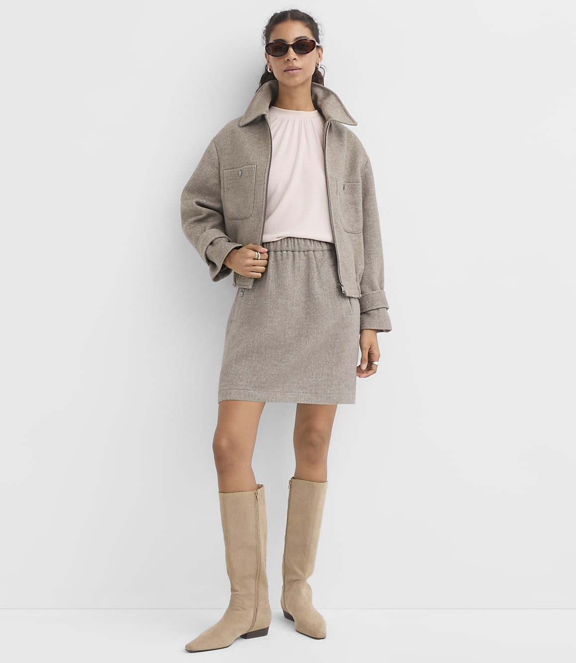 A woman in a light pink top, beige jacket, and matching skirt stands against a plain white background, wearing knee-high beige boots and sunglasses.