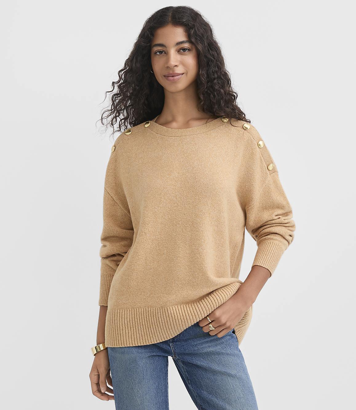A woman in a tan sweater with gold buttons on the shoulders and blue jeans stands against a plain white background.