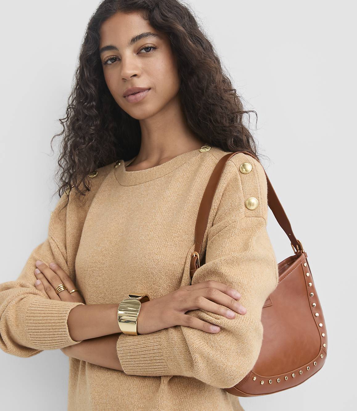 A woman in a tan sweater with gold buttons on the shoulder poses with a brown studded shoulder bag against a plain gray background.