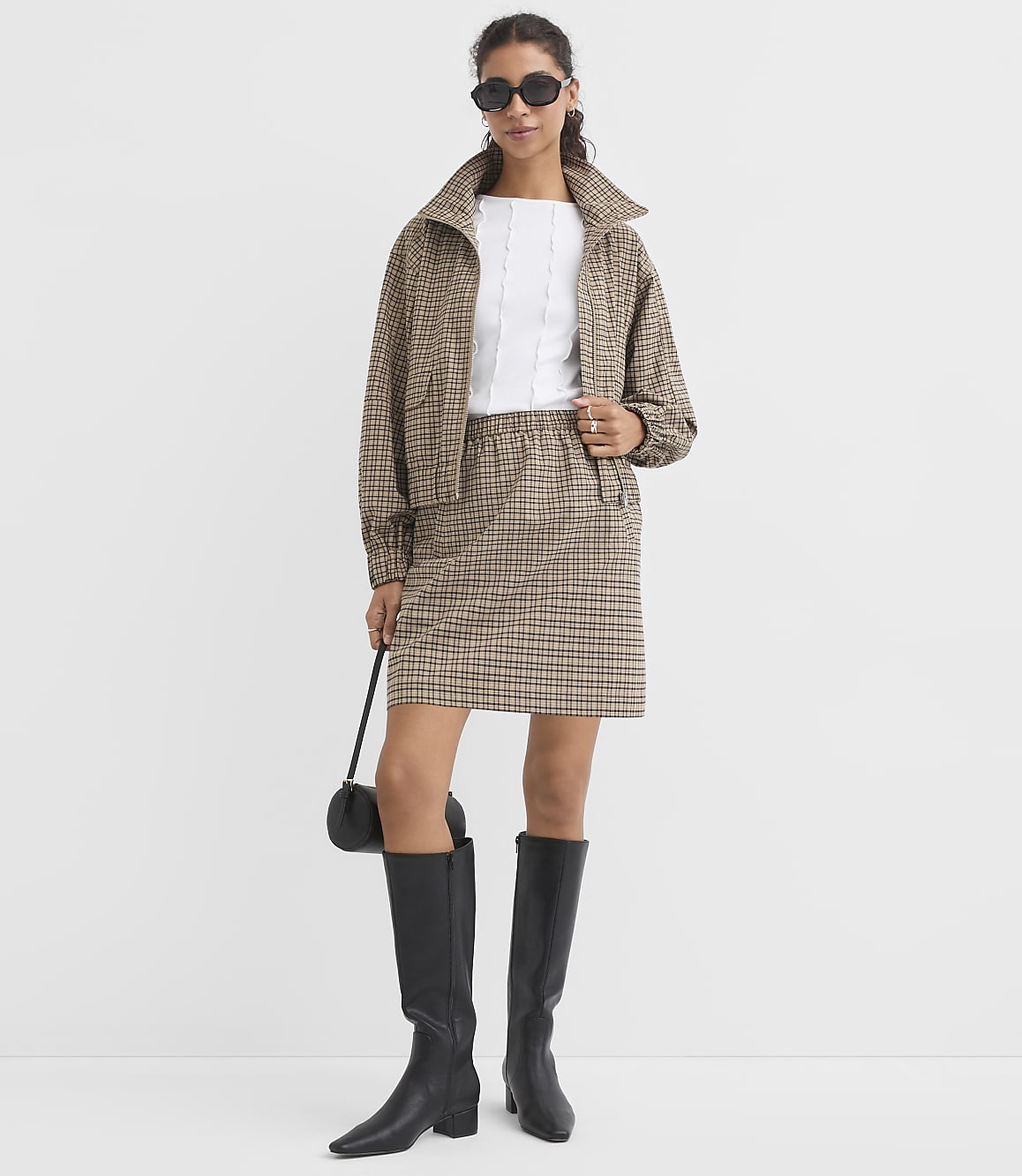 A woman in a plaid jacket and skirt stands against a plain white background, holding a black purse.