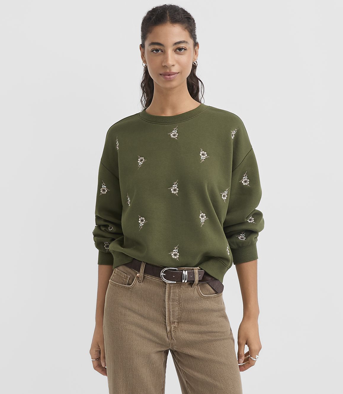 A woman in a green embroidered sweatshirt and brown pants stands against a plain gray background.