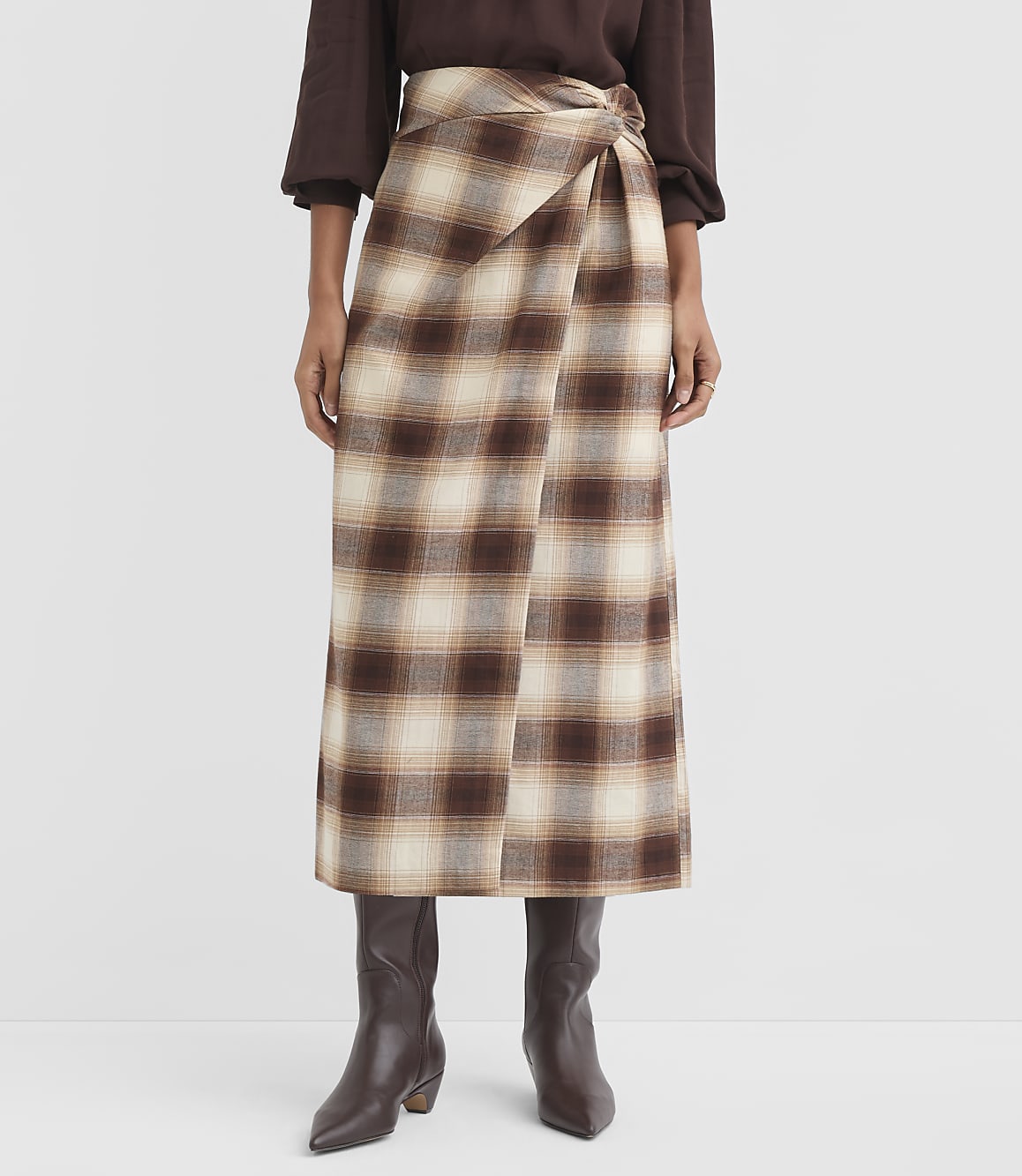 A person stands wearing a brown and cream plaid wrap skirt and dark brown boots against a plain white background.