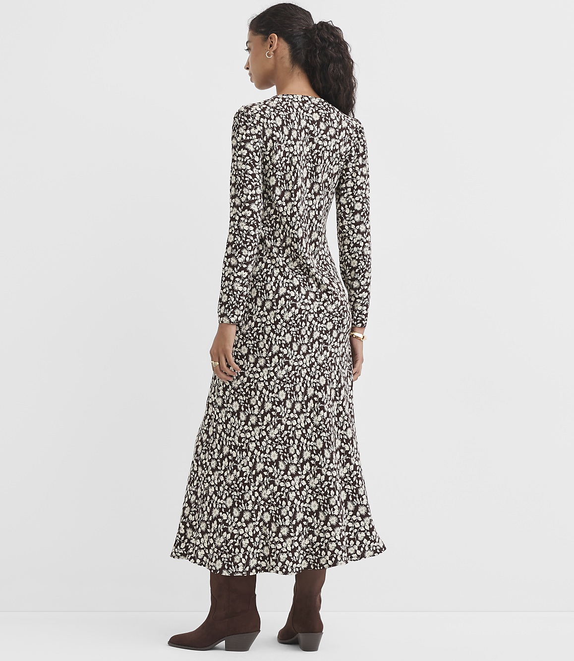 A woman in a long-sleeved, floral-print dress and brown boots stands against a plain, light gray background.