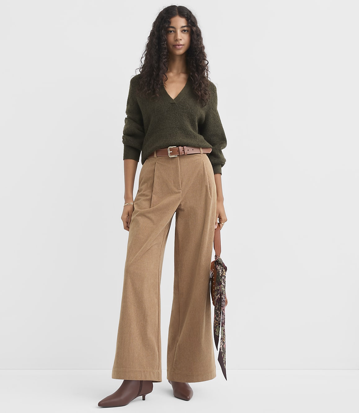 A woman in a green sweater and tan corduroy pants stands against a plain white background.