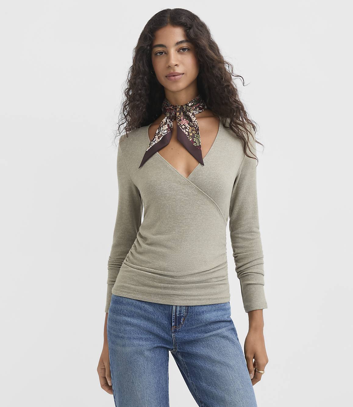 A woman in a light green wrap top and blue jeans stands against a plain gray background, a floral scarf tied around her neck.
