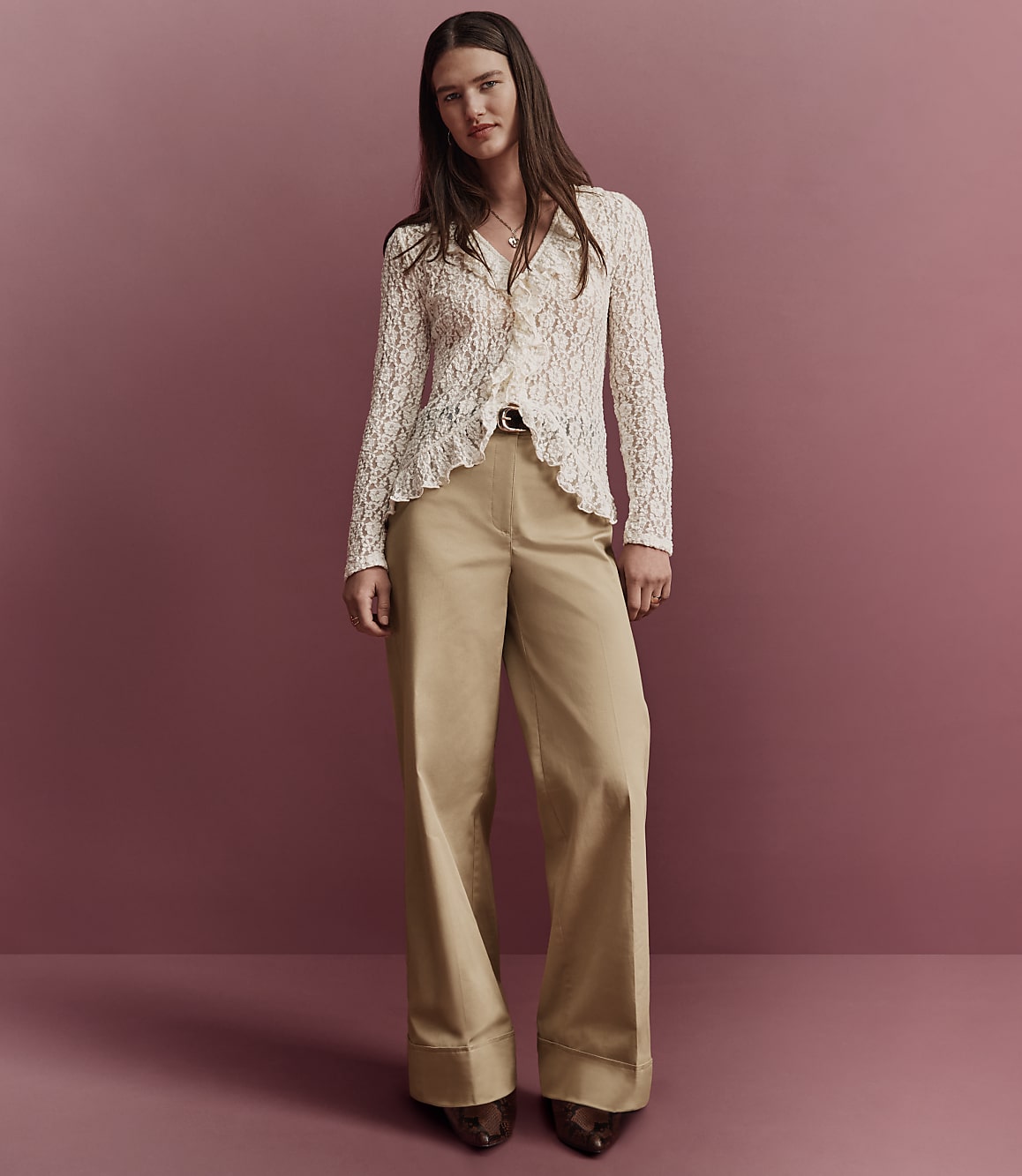 A woman in a white lace top and tan wide-leg pants stands against a pink background.