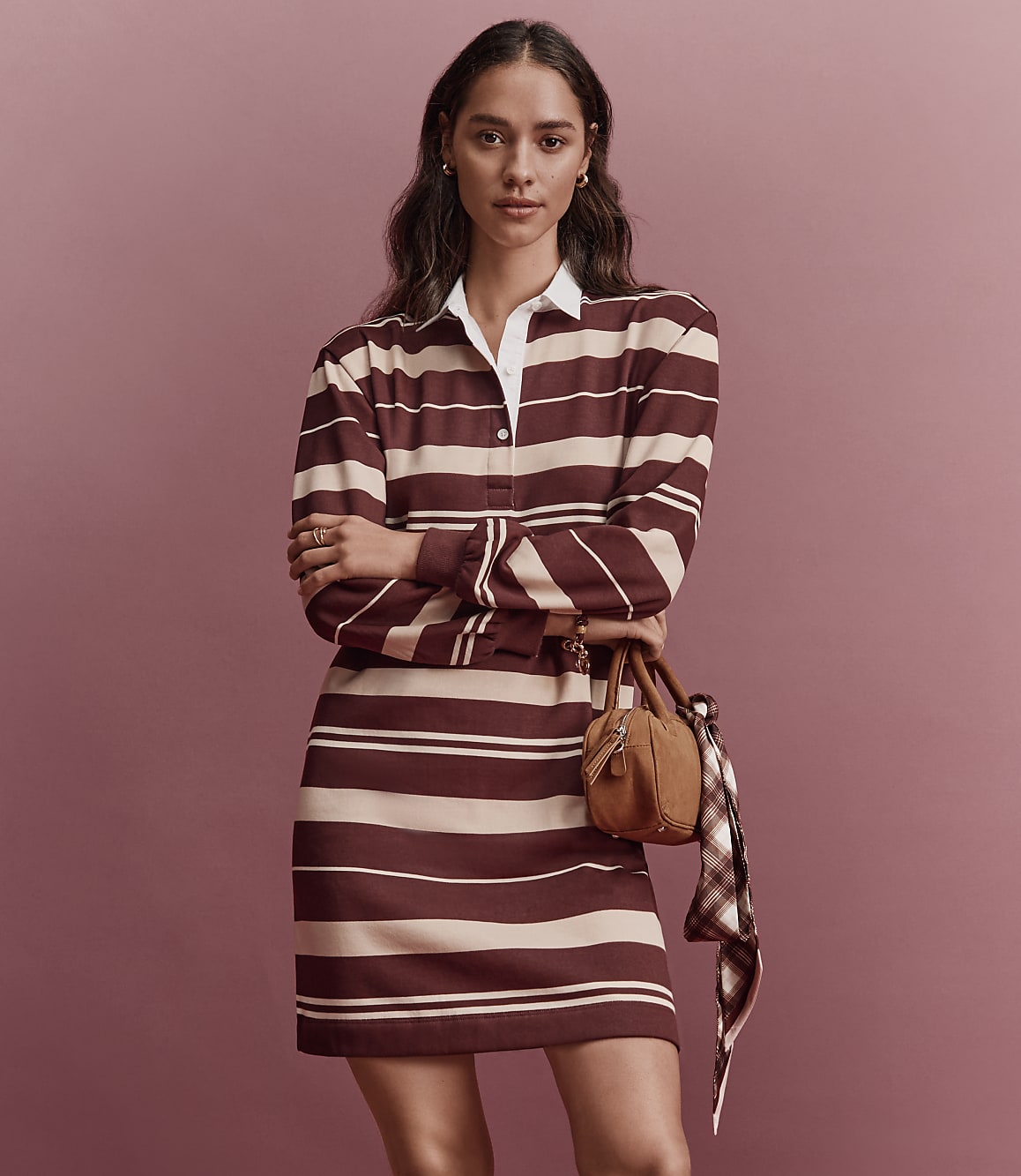A woman in a striped rugby dress stands with her arms crossed in front of a plain pink background, holding a small brown handbag with a scarf draped over it.