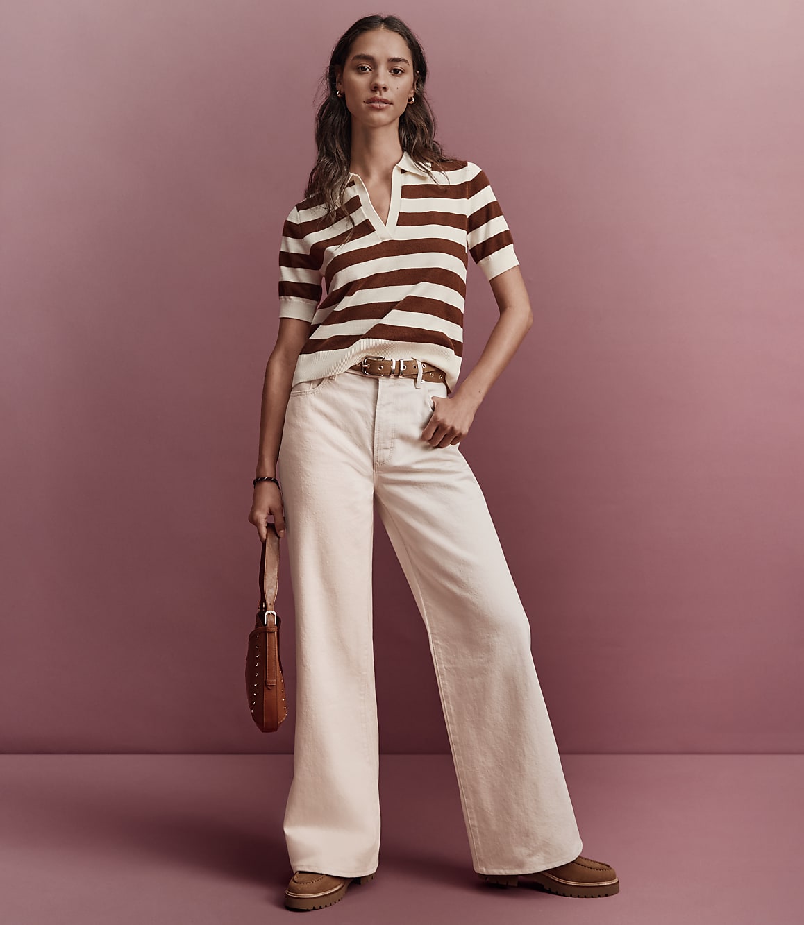 A woman in a striped polo shirt and wide-leg pants stands against a muted pink background.