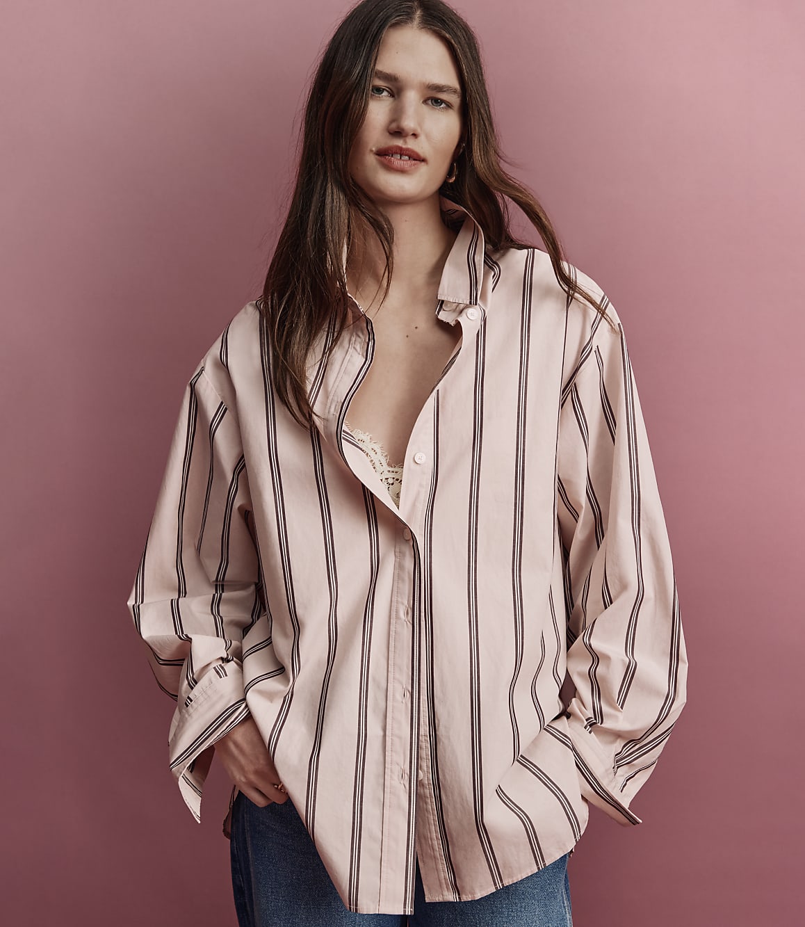 A woman in a pink and black striped oversized shirt and blue jeans stands against a plain pink background.