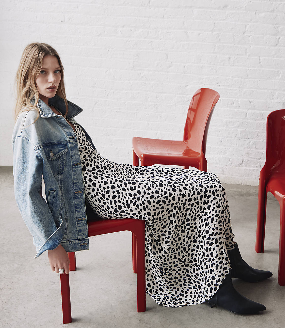 A young woman in a dalmatian print dress and denim jacket sits on a red chair in front of a white brick wall.