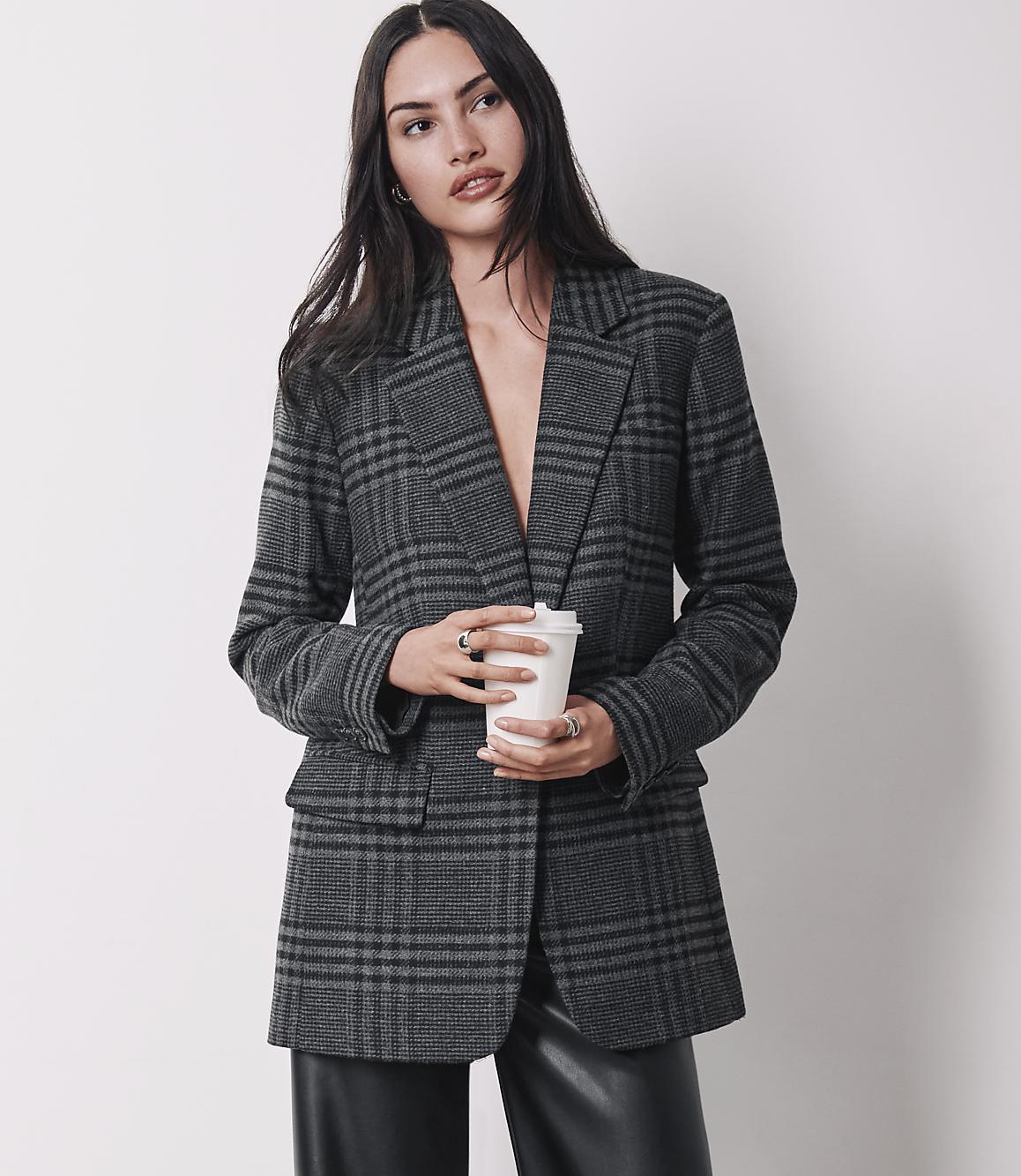 A woman in a plaid blazer and leather pants holds a coffee cup against a plain background.