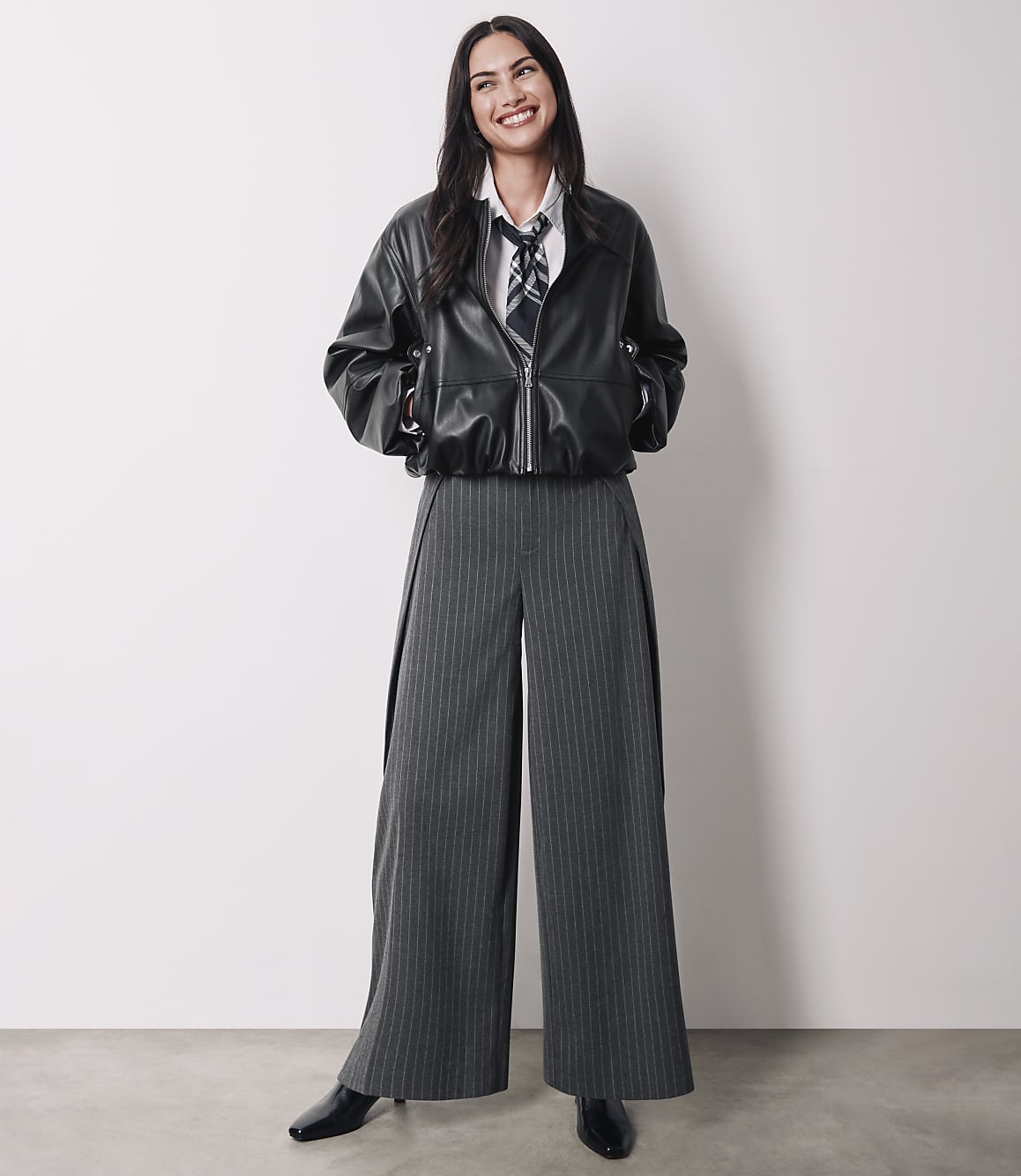 A smiling woman in a black leather jacket and pinstripe pants stands against a plain white background.