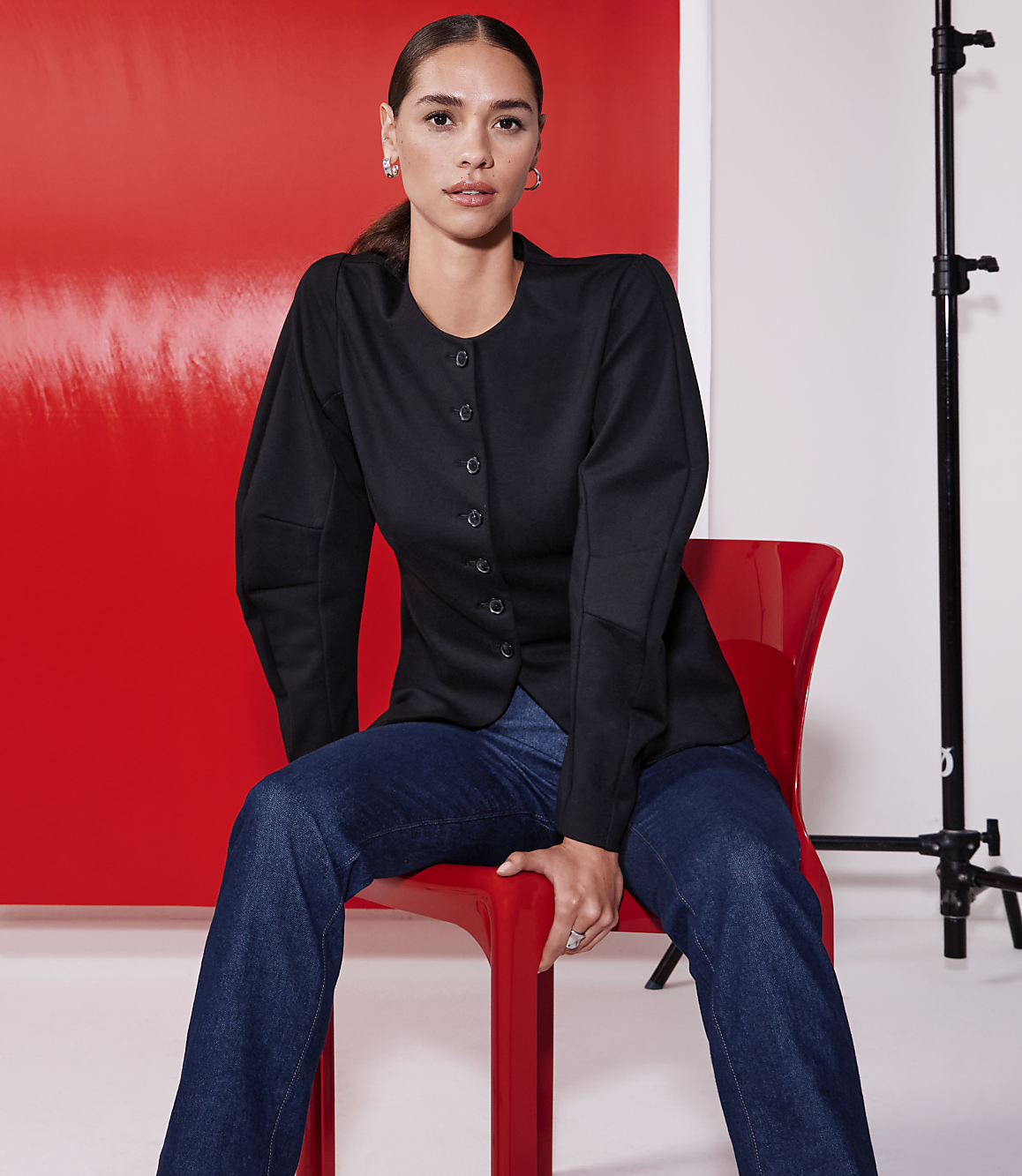 A woman in a black jacket and jeans sits on a red chair against a vibrant red backdrop with studio lighting equipment visible.