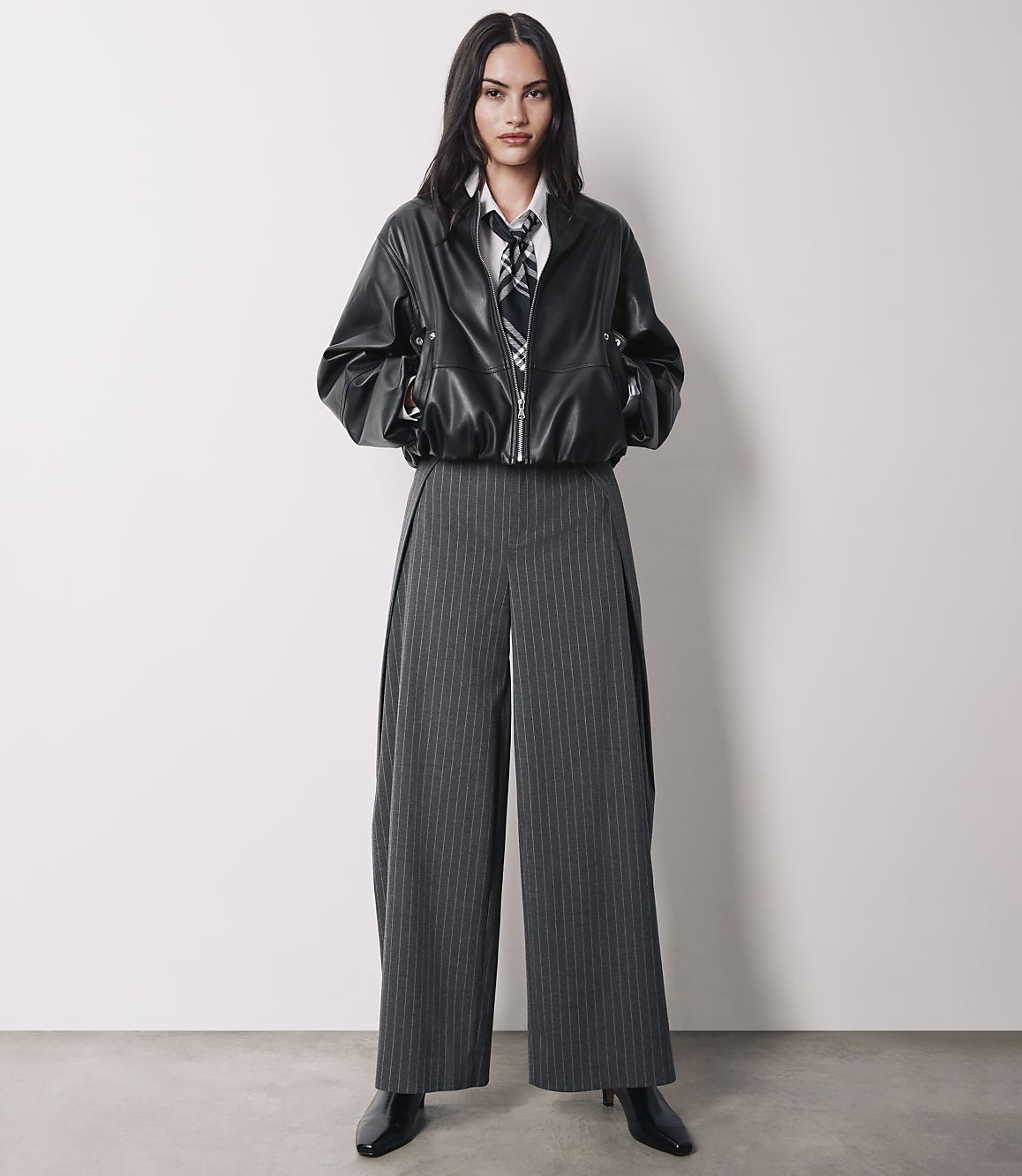 A woman in a black leather jacket and pinstripe pants stands against a plain white background.