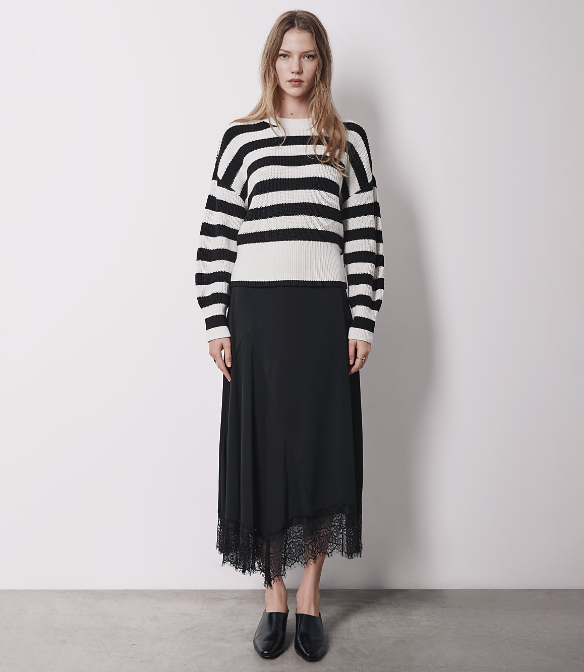 A woman in a striped sweater and black skirt stands against a plain white background.