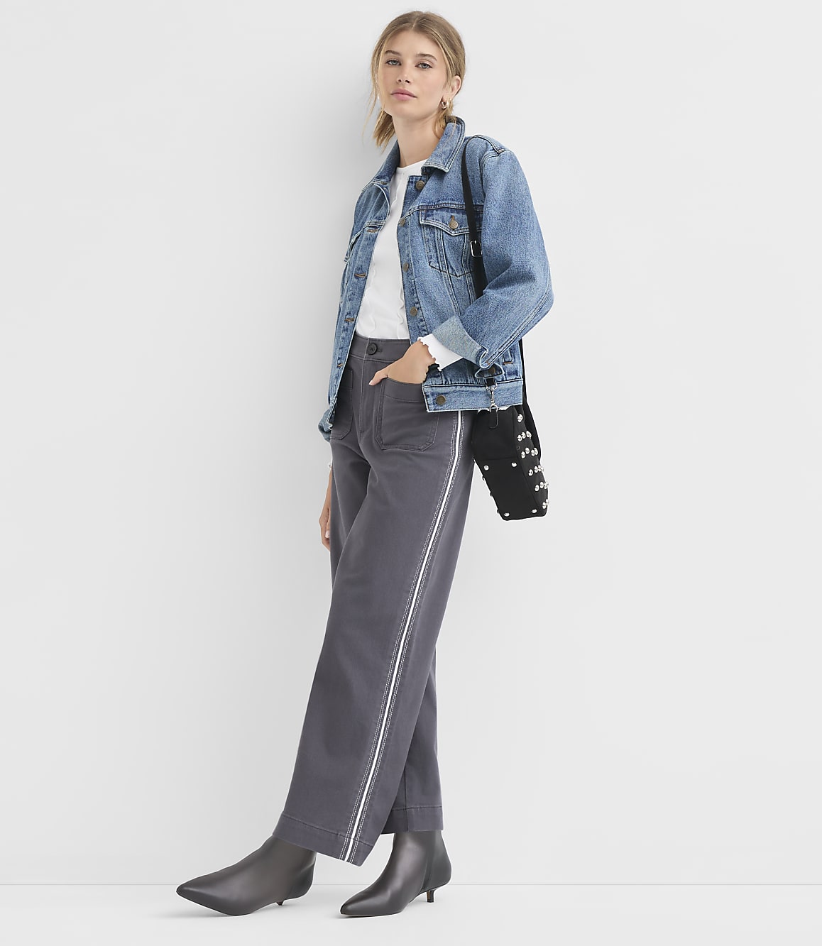 A woman in a denim jacket and gray pants stands against a plain white background.