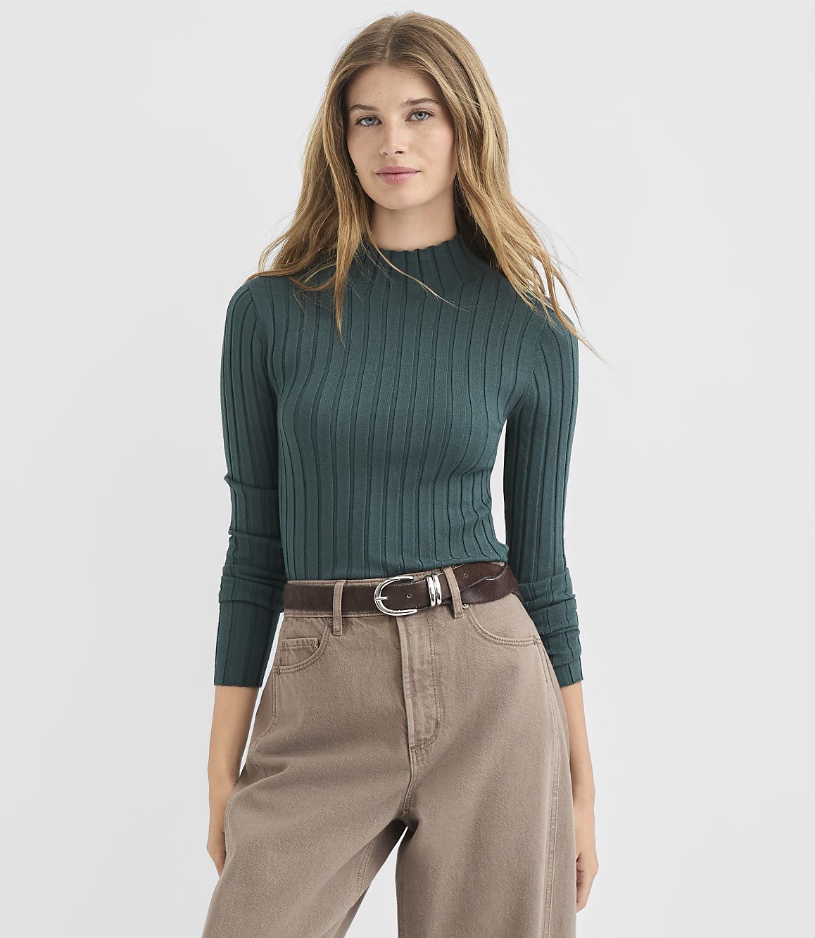 A woman in a teal ribbed sweater and brown pants stands against a plain white background.
