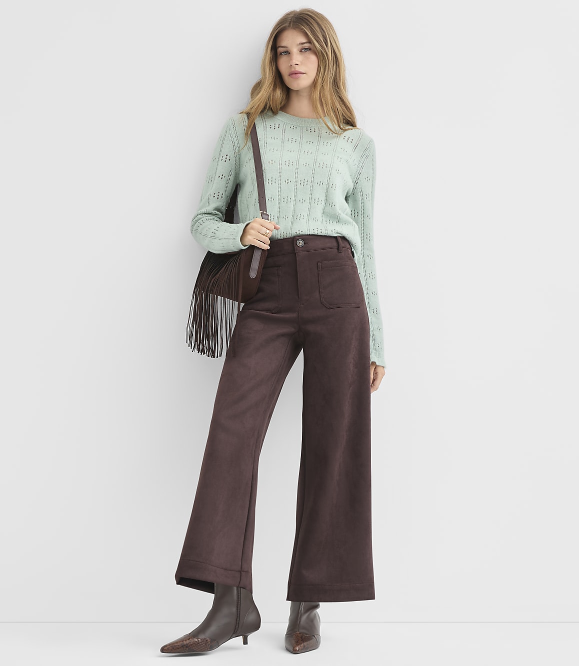 A woman in a light green sweater and brown wide-leg pants stands against a plain white background.