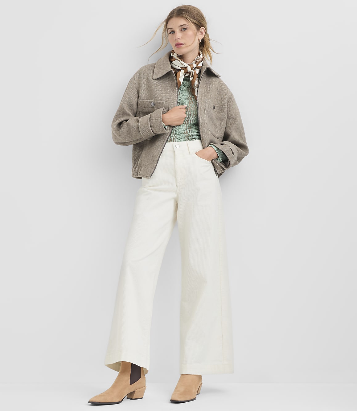 A woman in a beige jacket, green patterned shirt, and white wide-leg pants stands against a plain white background.