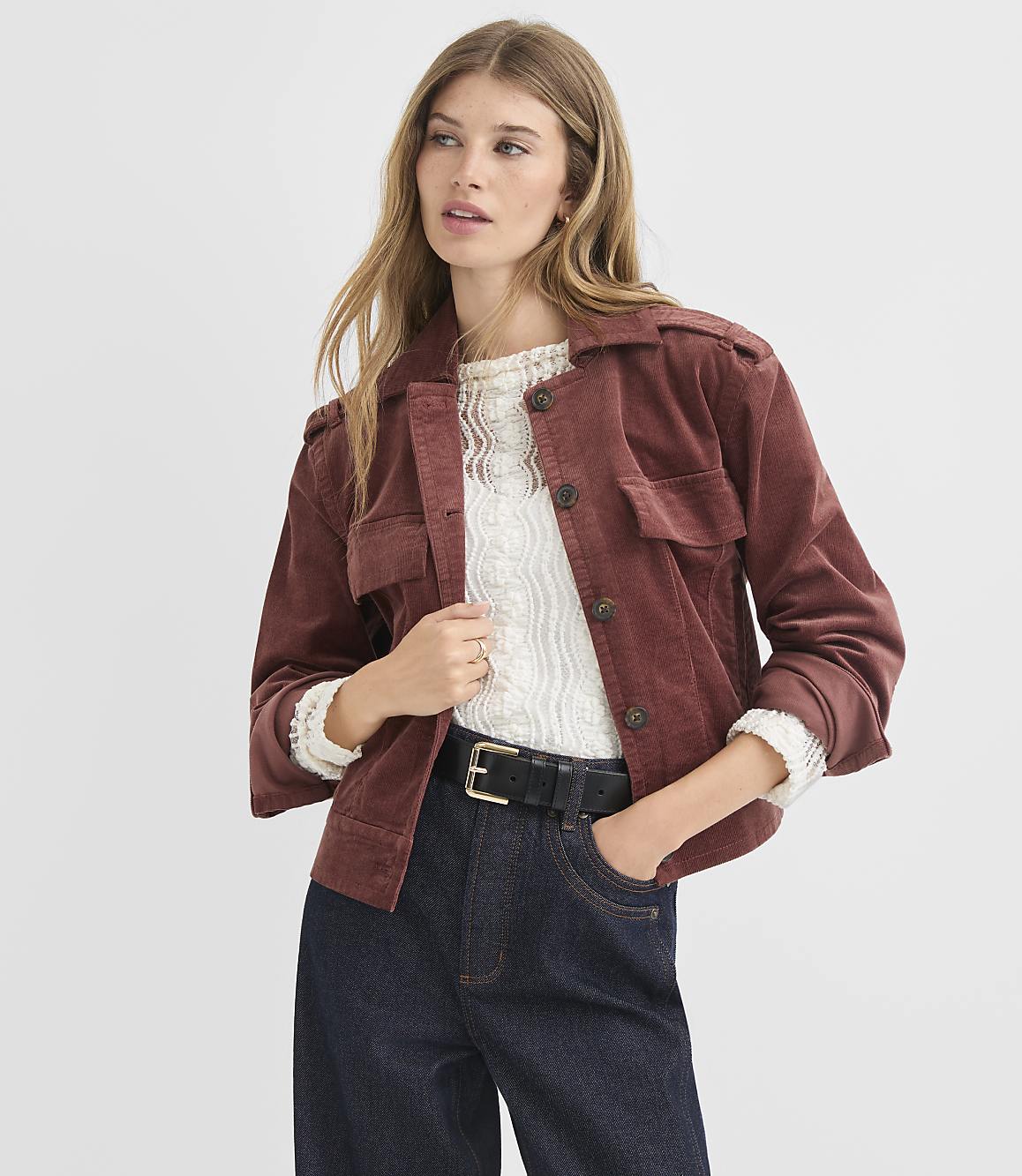 A woman in a rust-colored corduroy jacket and dark jeans stands against a plain white background.