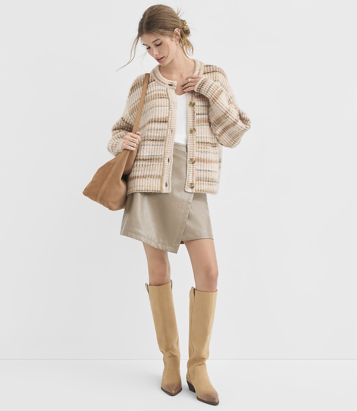 A woman in a plaid cardigan, white top, and tan skirt stands against a plain white background, accessorized with a brown tote bag and knee-high boots.