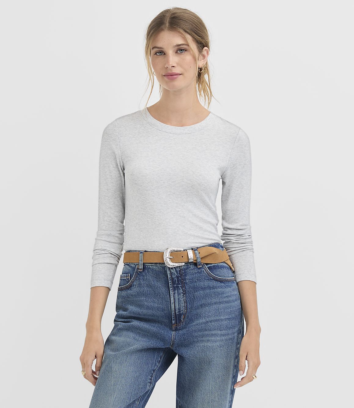A woman in a grey long-sleeved shirt and blue jeans with a brown belt stands against a plain white background.