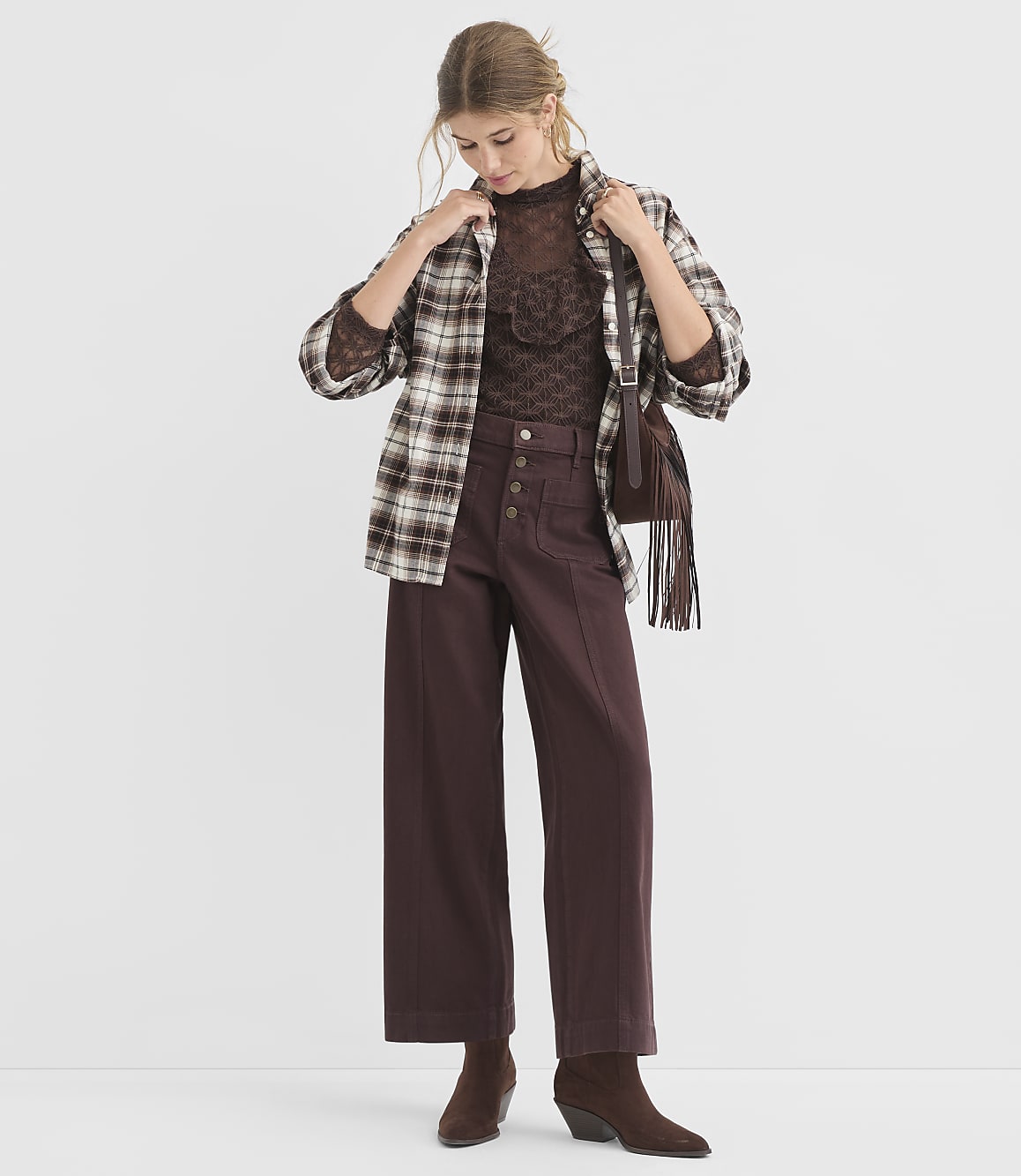 A woman in a brown plaid shirt and wide-leg pants stands against a plain white background.