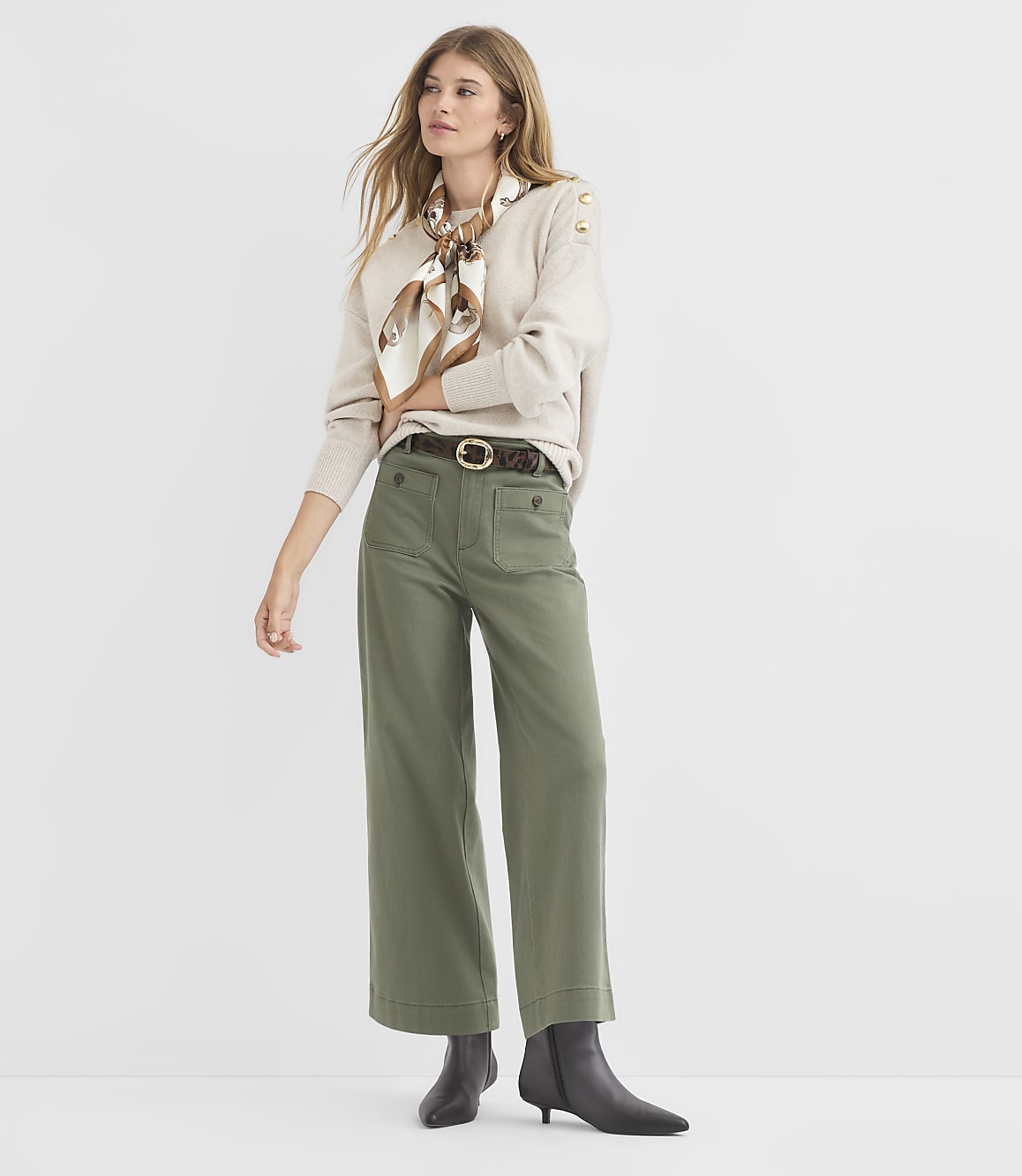 A woman in a beige sweater, patterned scarf, and olive green wide-leg pants stands against a plain white background.