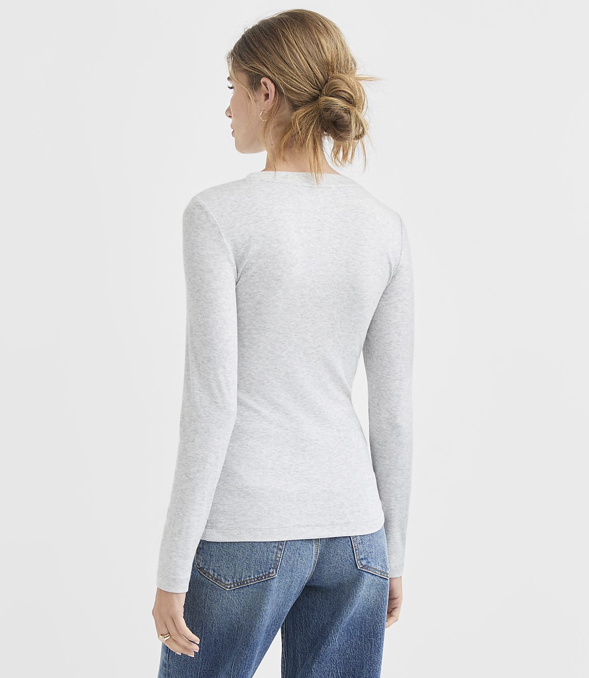 A woman in a grey long-sleeved shirt and blue jeans stands against a plain white background.