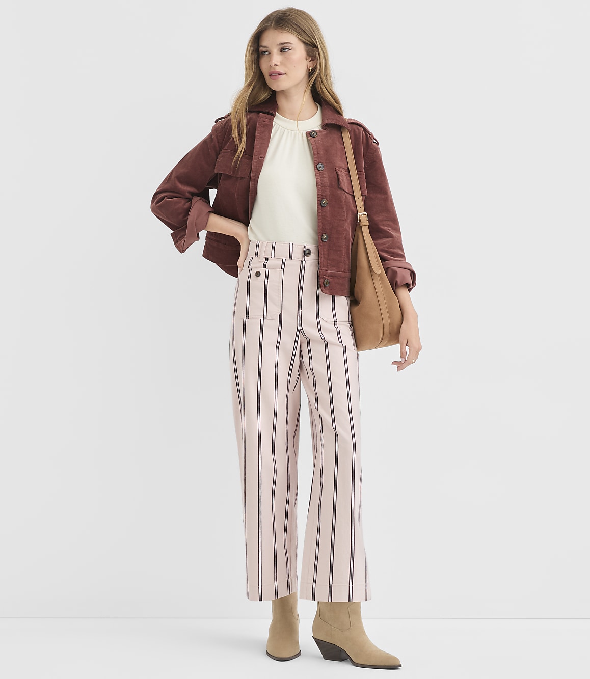 A woman in a maroon jacket and pink striped pants stands against a plain white background, holding a brown purse.