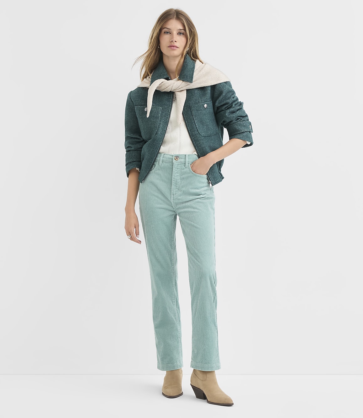 A woman in a teal jacket and light blue corduroy pants stands against a plain white background.