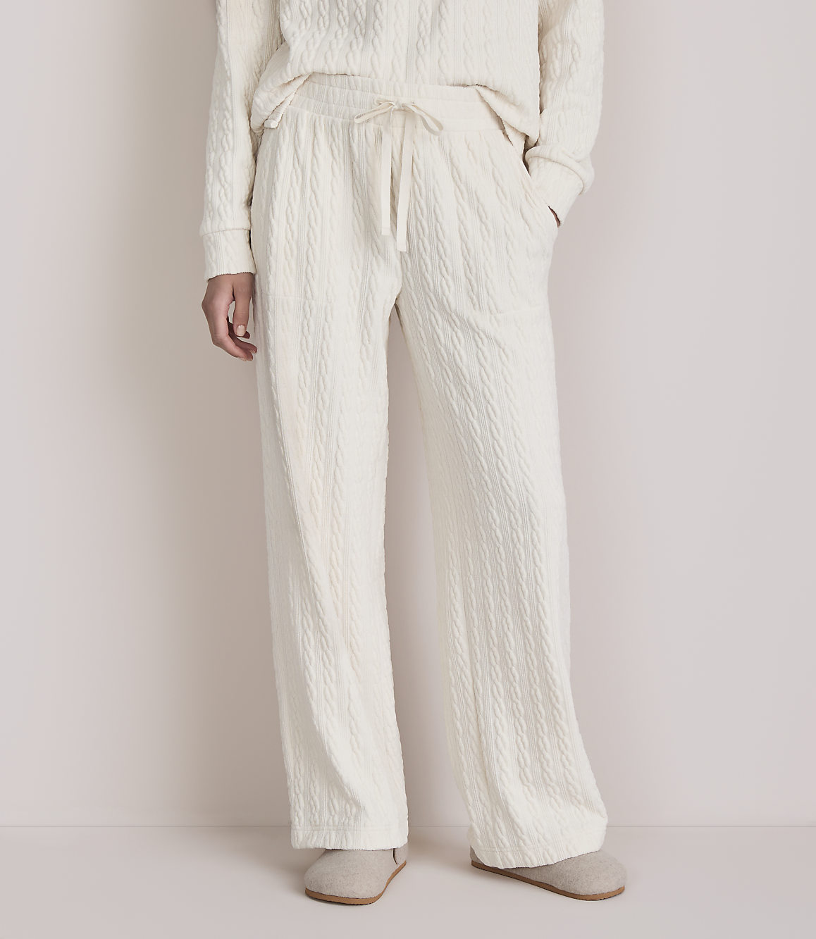 A person stands in cream-colored cable-knit pajamas and slippers against a plain, light-colored background.