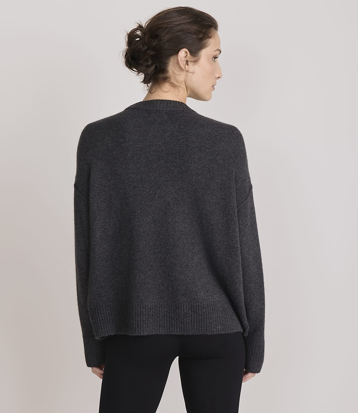 A woman in a dark grey sweater and black pants stands against a plain grey background.
