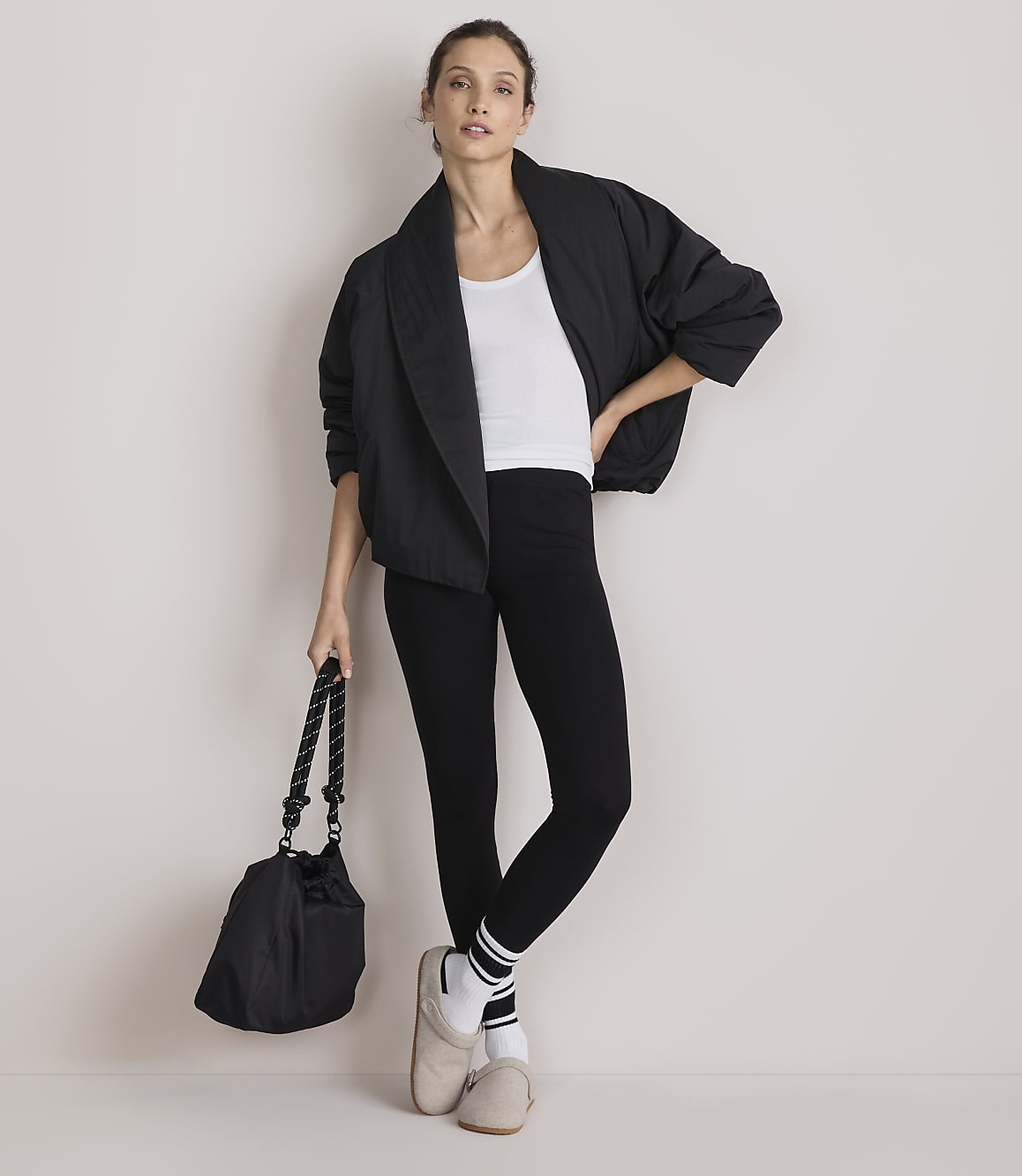 A woman in a black jacket, white tank top, and black leggings stands with a black bag in her left hand against a plain white background.