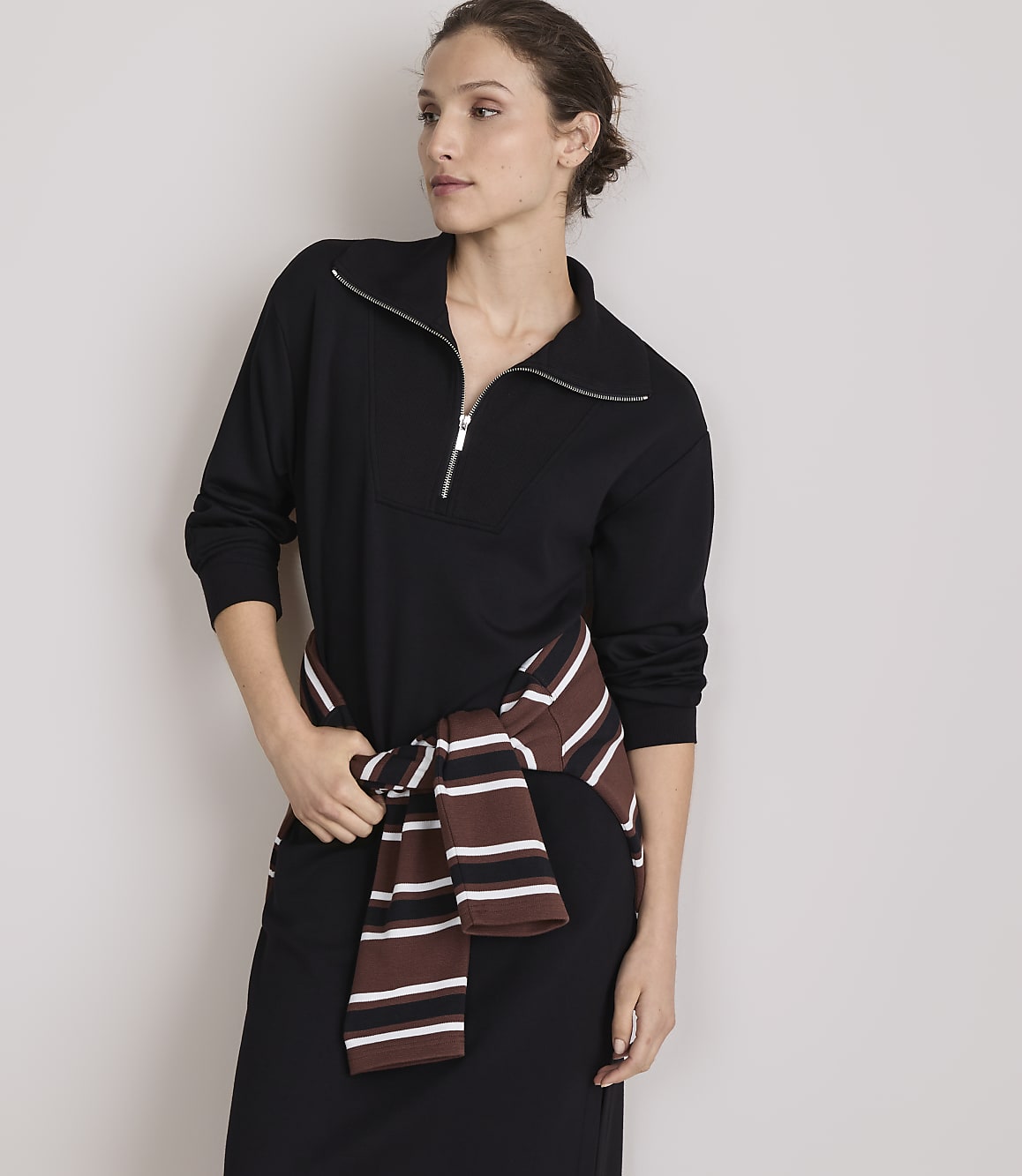 A woman in a black dress with a striped sweater tied around her waist stands against a plain gray background.