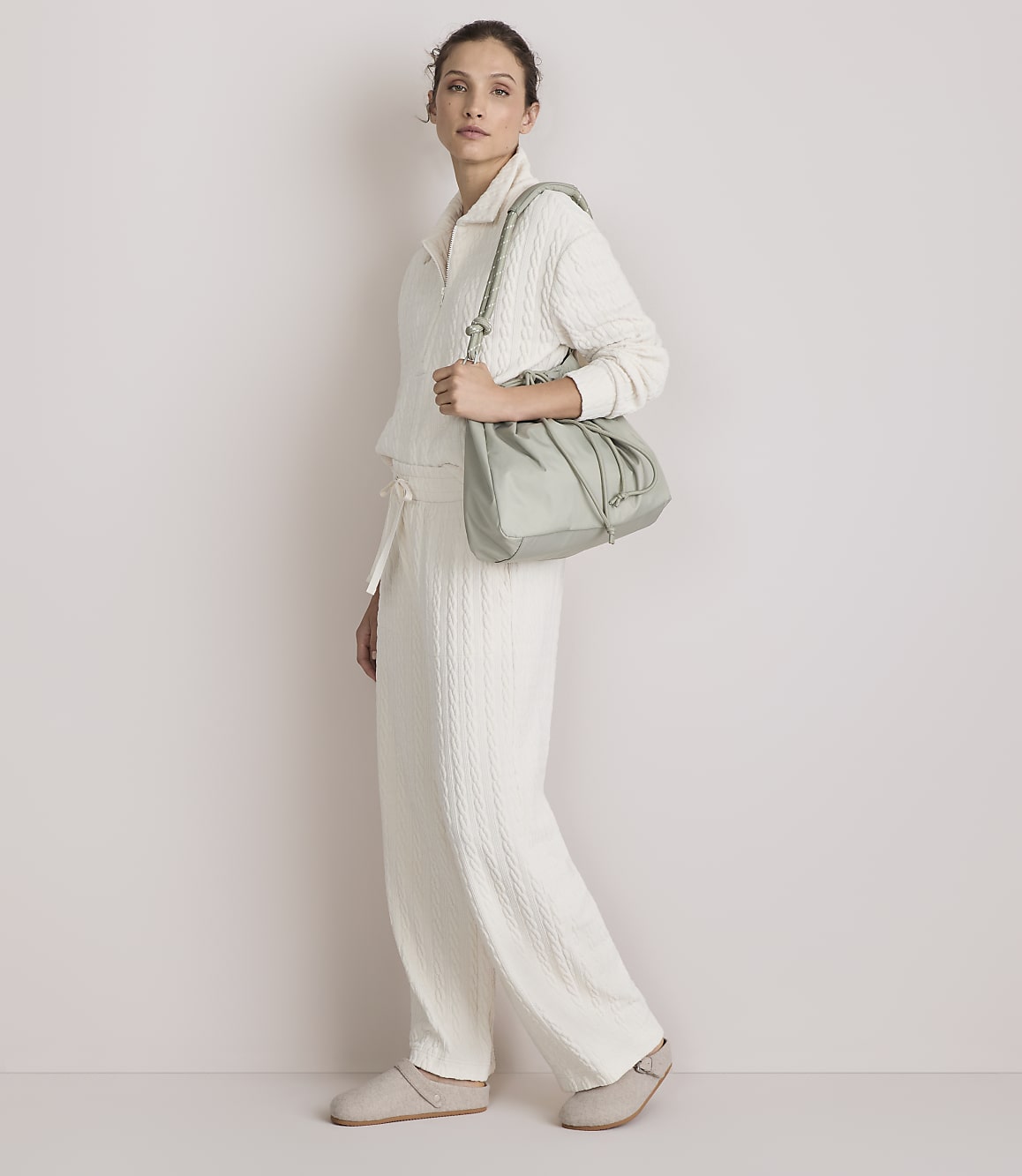 A woman in a cream-colored loungewear set and slippers walks with a light green drawstring bag against a plain, light-colored background.