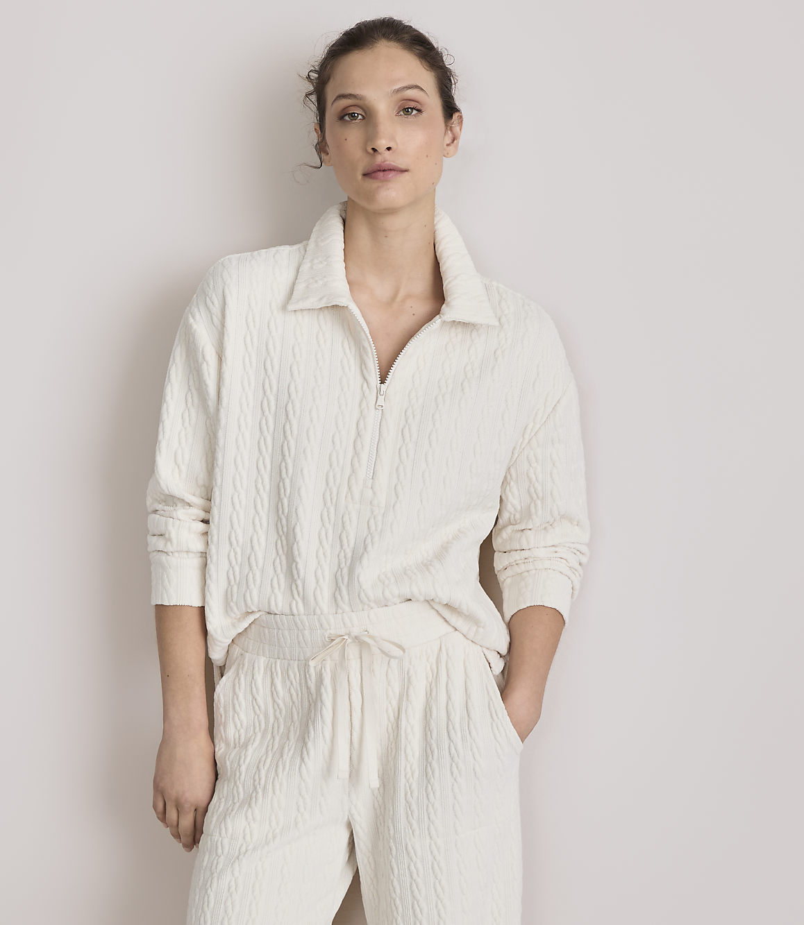 A woman in a cream-colored cable-knit loungewear set stands against a plain, light-colored wall.